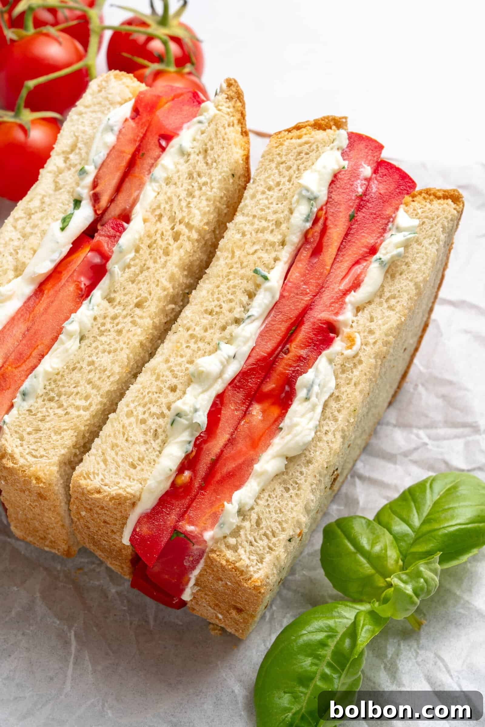 Two thick slices of sandwich bread filled with layers of fresh tomato and herbed cream cheese are placed on parchment paper, with vine tomatoes and fresh basil nearby.