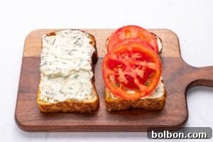 Two slices of bread on a wooden cutting board, one spread with herb cream cheese and the other topped with cream cheese and sliced tomatoes—perfect for trying out your next Tomato Sandwich Recipe.
