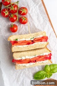 A Tomato Sandwich Recipe features thick tomato slices and a creamy filling, cut in half and stacked on parchment paper. Fresh basil leaves and a cluster of ripe tomatoes rest nearby on a wooden board.