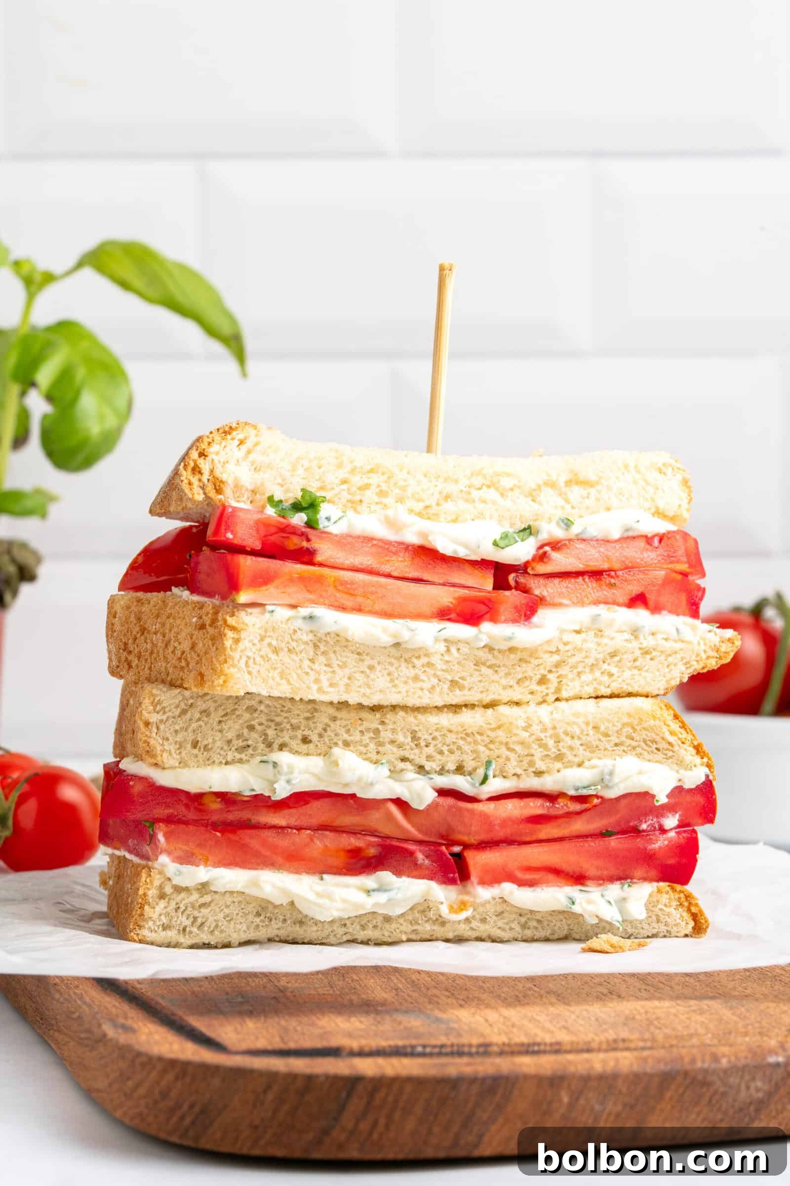 A stack of two tomato sandwiches with mayonnaise and herbs, made with thick slices of white bread, is shown on a wooden board. A skewer holds the sandwiches together. Fresh tomatoes and basil are in the background.