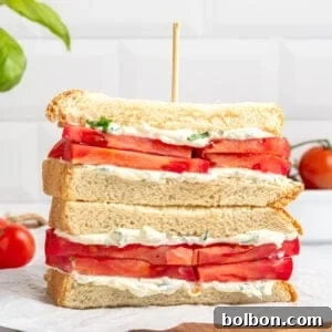 A stacked tomato sandwich with thick tomato slices, creamy spread, and herbs on white bread, held together with a skewer, sits on parchment paper atop a wooden board. Try this classic Tomato Sandwich Recipe—fresh tomatoes and basil complete the scene.