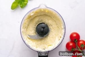 A food processor bowl filled with creamy ricotta cheese mixture, perfect for your next Tomato Sandwich Recipe, sits beside cherry tomatoes and fresh basil leaves on a white surface.
