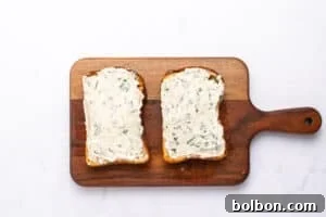 Two slices of toasted bread with a creamy white herb spread sit side by side on a wooden cutting board—a perfect start to any Tomato Sandwich Recipe—set against a clean white background.