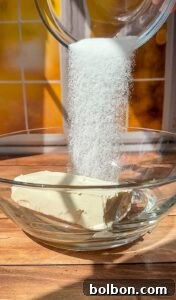 Decadent Pumpkin Bread with Cream Cheese 5 Granulated sugar is being poured from a glass bowl onto a block of cream cheese in a clear glass mixing bowl, set on a wooden surface with a sunny window in the background.