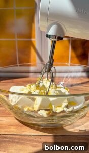 Decadent Pumpkin Bread with Cream Cheese 6 A hand mixer with metal beaters is positioned above a glass bowl containing cream cheese and butter, ready to be blended. Warm sunlight streams through a window in the background.