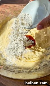 Decadent Pumpkin Bread with Cream Cheese 7 A hand pours flour from a small white bowl into a glass mixing bowl filled with creamy batter, ready to be mixed together.