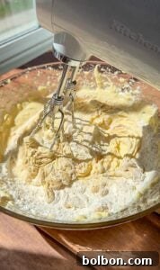 Decadent Pumpkin Bread with Cream Cheese 8 A hand mixer blending flour into a creamy batter in a glass bowl on a wooden surface, with sunlight streaming in from a window nearby.