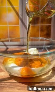 Decadent Pumpkin Bread with Cream Cheese 9 A glass measuring cup pours oil into a glass bowl containing eggs, sugar, and a dollop of cream, with sunlight shining through a window in the background.