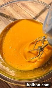 Decadent Pumpkin Bread with Cream Cheese 10 A close-up of an electric mixer blending a thick, bright orange batter in a clear glass bowl placed on a wooden surface.