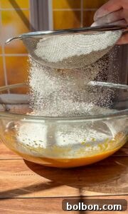 Decadent Pumpkin Bread with Cream Cheese 11 A close-up of a hand sifting flour through a metal sieve into a glass bowl containing a yellow batter, with sunlight streaming through a window and yellow tiles in the background.