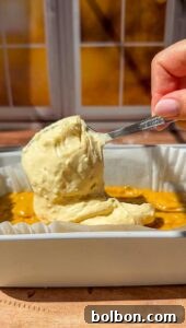 Decadent Pumpkin Bread with Cream Cheese 13 A hand holds a spoonful of thick, creamy batter over a rectangular baking dish with a yellow mixture inside, set on a wooden surface near a sunlit window.