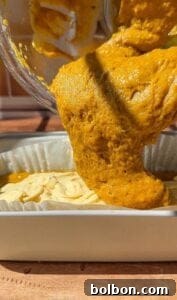 Decadent Pumpkin Bread with Cream Cheese 14 Thick, golden-orange batter is being poured from a bowl into a parchment-lined baking dish, ready to be baked. The texture is airy and slightly bubbly, with sunlight highlighting the rich color.