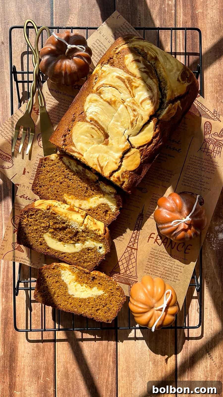 Decadent Pumpkin Bread with Cream Cheese 3 A loaf of pumpkin bread with a cream cheese swirl is sliced on parchment paper atop a cooling rack. Two pumpkin-shaped decorations, a gold fork, and knife are nearby, all on a wooden table in sunlight.