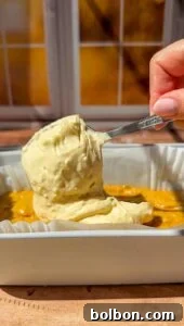Decadent Pumpkin Bread with Cream Cheese 15 A hand holds a spoonful of thick, creamy batter over a rectangular baking dish with a yellow mixture inside, set on a wooden surface near a sunlit window.