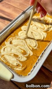 Decadent Pumpkin Bread with Cream Cheese 16 A hand swirls cream cheese into pumpkin batter with a skewer in a loaf pan, creating a marbled effect before baking. The pan sits on a wooden surface.