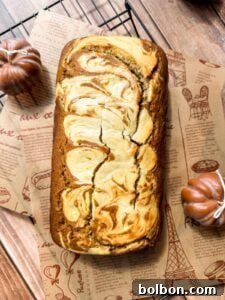 Decadent Pumpkin Bread with Cream Cheese 17 A loaf of Pumpkin Bread with Cream Cheese Filling, topped with a creamy swirl, sits on patterned parchment paper, flanked by two small decorative pumpkins atop a wooden surface.