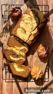 Decadent Pumpkin Bread with Cream Cheese 18 A marble pumpkin bread loaf with cream cheese swirls is sliced on a cooling rack, surrounded by decorative mini pumpkins and gold utensils, with warm sunlight casting soft shadows.