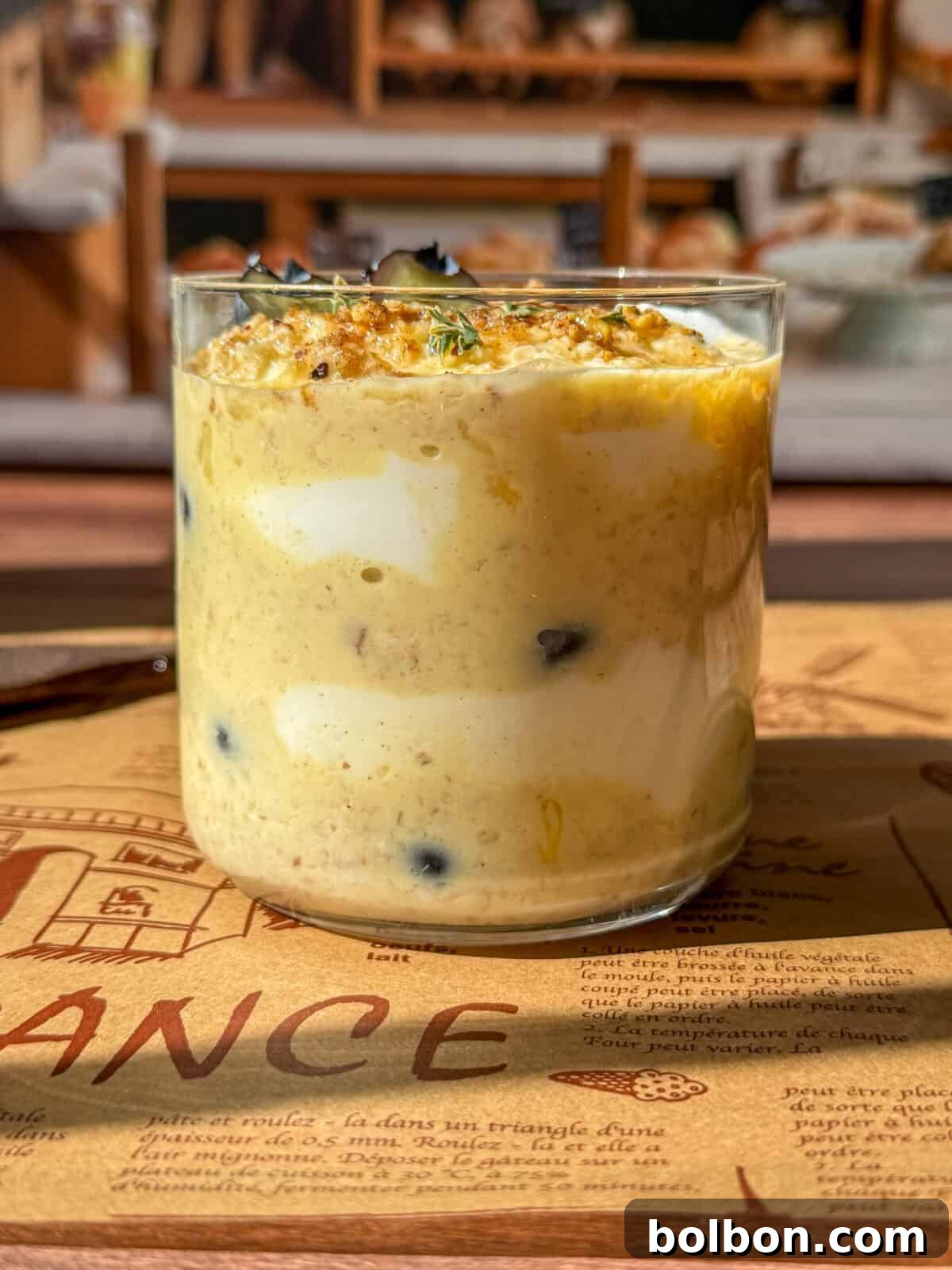 A elegant glass jar filled with layers of creamy overnight oats, meticulously topped with golden granola and fresh herbs, positioned on a decorative placemat with the soft, blurred background of a bakery interior, evoking a sense of calm morning luxury.