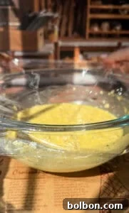 A clear glass bowl containing a yellow batter is securely covered with plastic wrap, resting on a table in a softly lit kitchen environment. A printed recipe sheet lies beneath the bowl, and the background subtly blurs into shelves and various kitchen implements, creating a warm, domestic scene.