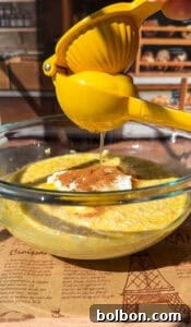 A hand skillfully squeezes a bright yellow lemon using a lemon squeezer, directing the fresh juice into a clear glass bowl. The bowl holds a creamy mixture of oats, yogurt, and a sprinkle of cinnamon, all illuminated by natural sunlight, with a bakery setting subtly in the background.