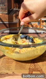 A hand holds a spoon, poised over a transparent glass bowl of creamy oatmeal, which is delicately adorned with fresh blueberries, finely grated apple, and a light dusting of cinnamon. The bowl rests on a decorative paper featuring illustrations of the Eiffel Tower, suggesting a charming European breakfast setting.