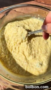 A hand holds a spoon, scooping a generous portion of a thick, yellowish, and visibly grainy creamy mixture from a glass bowl. The scene is brightly lit by natural sunlight, highlighting the rich texture of the prepared dish.