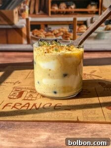 A clear glass jar brimming with a creamy, layered dessert is beautifully topped with fresh blueberries and a scattering of crumbs. The jar rests on a warm brown placemat adorned with the word 