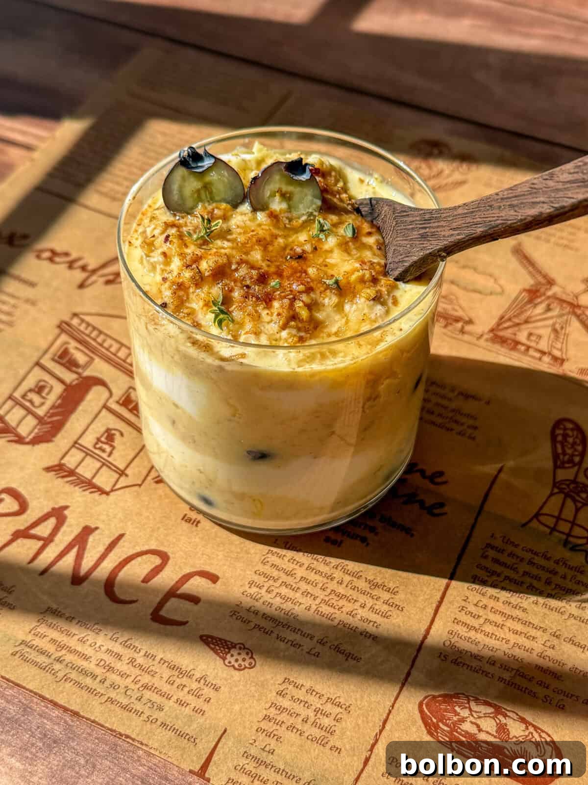 An elegantly styled glass of creamy dessert, garnished with two fresh blueberries, a delicate sprinkle of caramelized sugar, and a sprig of fresh herbs. It rests on a rustic wooden table alongside a French-themed placemat. A wooden spoon is gently placed within the dessert, and warm sunlight streams in, casting soft shadows across the inviting scene.
