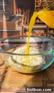 A glass jug pours vibrant orange juice into a glass bowl brimming with rolled oats, positioned on a rustic wooden table. In the softly blurred background, an inviting display of fresh bakery goods and artisanal bread hints at a delicious morning ahead.