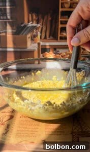 A hand holds a whisk, diligently mixing oats and a golden liquid within a clear glass bowl, set upon a warm wooden table. The backdrop reveals a charming, blurred bakery display featuring various breads and pastries, suggesting a cozy and culinary ambiance.