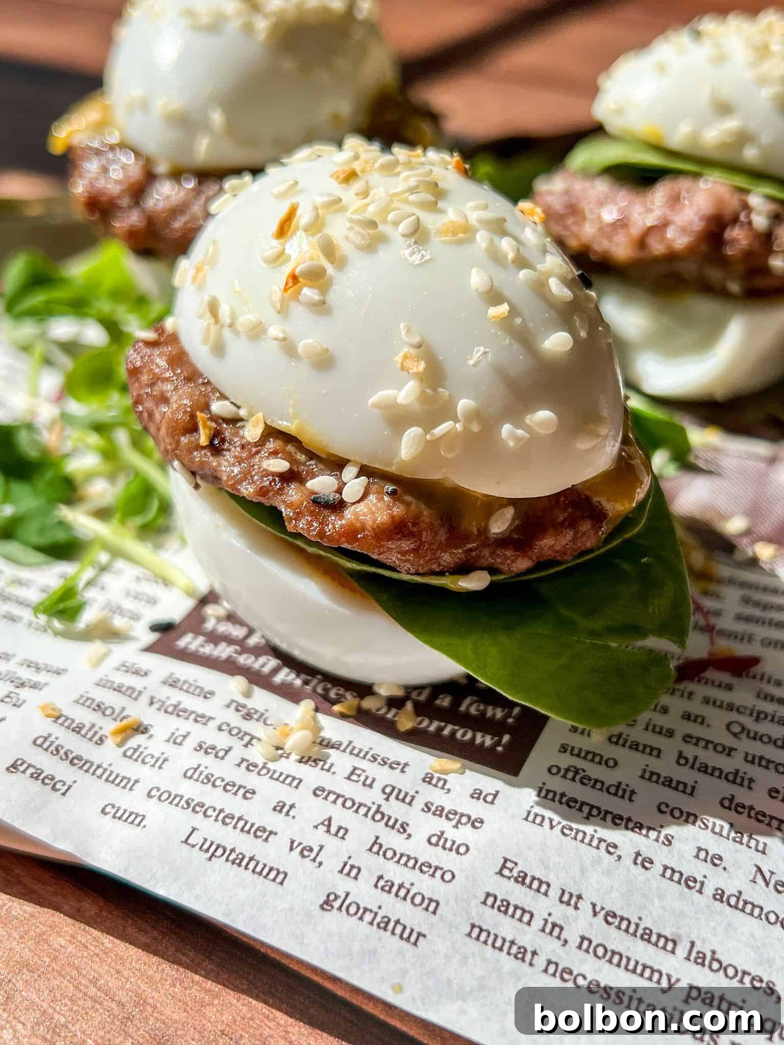 Close-up of Mini Egg Burgers—sliders made with boiled egg halves as buns, filled with a small beef patty, spinach leaves, and sesame seeds sprinkled on top—all placed on a sheet of printed newspaper.