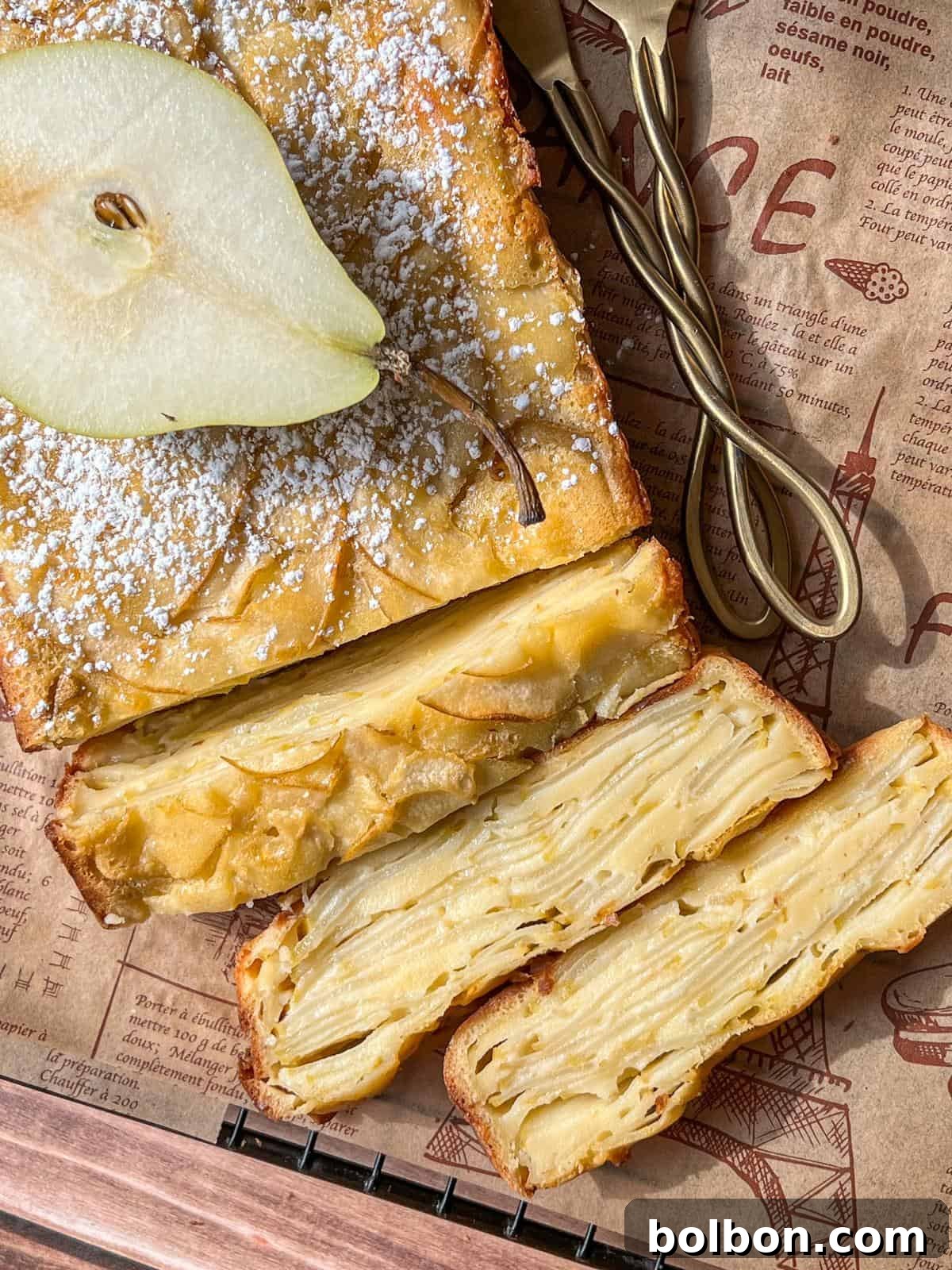 A perfectly baked Invisible Pear Cake, lightly dusted with powdered sugar, sits on parchment paper. A fresh pear slice garnishes the top, and a cross-section reveals golden, intricate layers of thinly sliced pears within the cake, ready to be enjoyed.