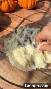 A hand mixes a yellow and white powdered mixture in a glass bowl on a wooden surface, with small orange pumpkins in the background—prepping ingredients for an Invisible Pear Cake.