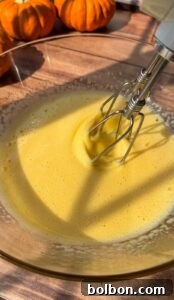 A close-up of an electric mixer blending yellow Invisible Pear Cake batter in a glass bowl on a wooden surface, with small pumpkins in the background and sunlight casting shadows across the scene.