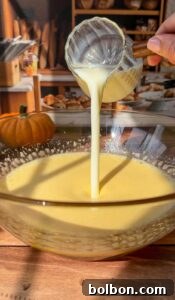 A hand pours creamy liquid from a glass measuring cup into a large bowl of yellow batter on a wooden table, preparing an Invisible Pear Cake, with baked goods and a pumpkin in the kitchen background.