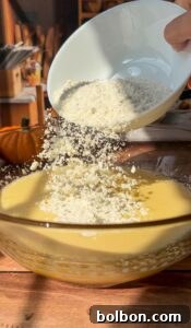 A hand pours flour from a white bowl into a glass mixing bowl of yellow batter. Sunlight streams onto the wooden table, hinting at an Invisible Pear Cake in progress, while a pumpkin sits blurred in the background.