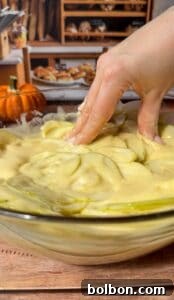 A hand mixes thinly sliced green apples in batter for an Invisible Pear Cake inside a glass bowl, with a small pumpkin and bakery shelves filled with bread visible in the background.