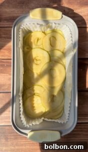 A rectangular baking pan lined with parchment paper holds thinly sliced green apples, arranged in overlapping layers for an Invisible Pear Cake. Sunlight casts gentle shadows on the wooden surface beneath the pan.
