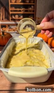 A hand pours creamy batter from a glass cup over thinly sliced apples arranged in a baking dish, creating an Invisible Pear Cake. Small pumpkins and bread are visible in the warm, autumn-themed background.