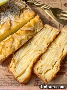 Close-up of a sliced cake with visible layers of thin apple slices, resembling the popular Invisible Pear Cake. The dessert sits on patterned parchment paper, with a fork and cake server in the background.