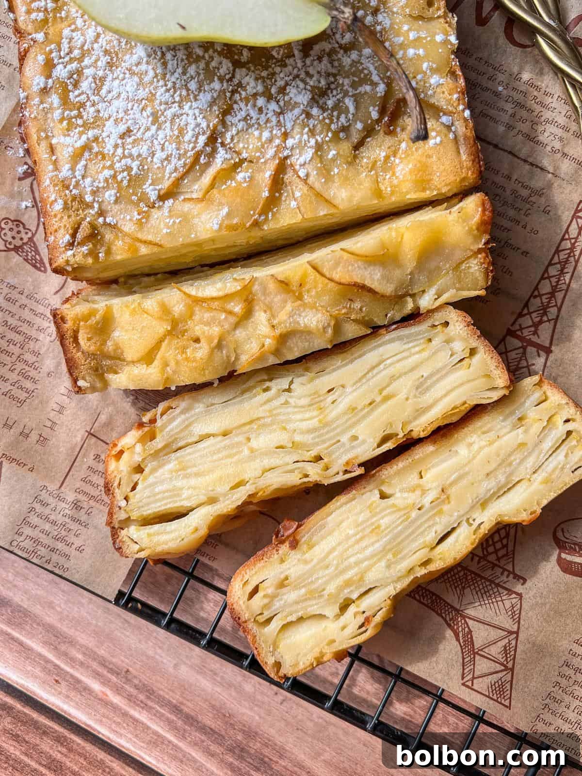 A close-up view of a slice of Invisible Pear Cake, elegantly dusted with powdered sugar. The intricate, golden-brown layers of thinly sliced pears are clearly visible, showcasing the cake's unique texture. It rests on a piece of printed parchment paper, positioned on a wire cooling rack over a rustic wooden surface.