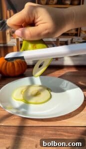 A hand slices a green pear using a white mandoline slicer over a white plate, preparing thin pear slices for Invisible Pear Cake. A small pumpkin sits in the background on a wooden surface.