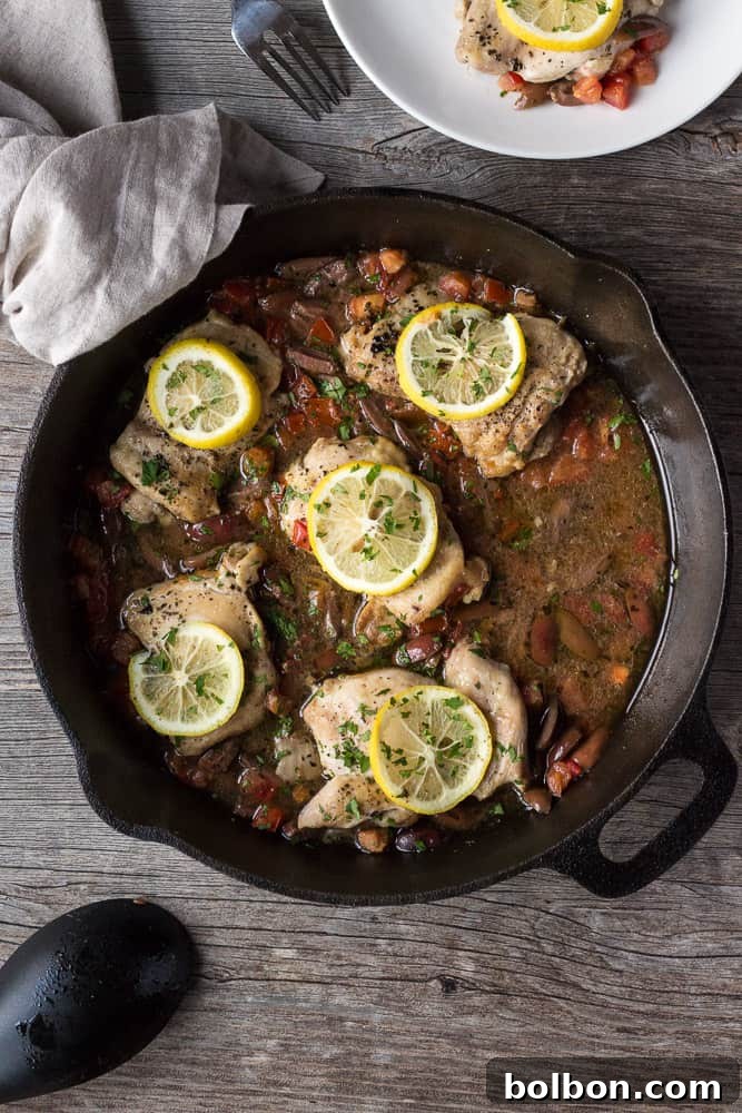 Greek Chicken Thighs recipe cooking in cast iron skillet, with lemon, Kalamata olives, and tomatoes. One pan dinner, quick and easy meal.