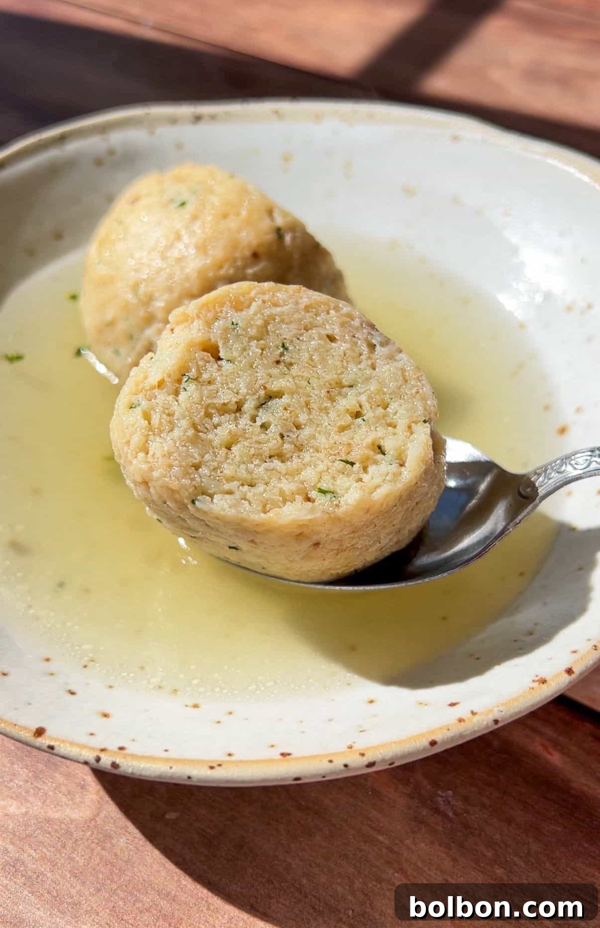 A bowl of clear broth with two sliced matzo balls, one resting on a spoon, sits on a wooden surface in bright sunlight—capturing the essence of Authentic Matzo Ball Soup.