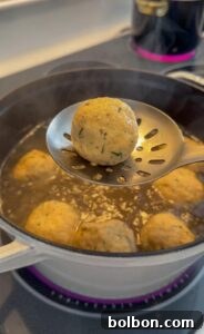 A slotted spoon holds a matzo ball over a pot of simmering broth, with more matzo balls cooking in the liquid below on a stovetop. Steam rises from the pot, capturing the warmth of authentic matzo ball soup, hot and freshly cooked.