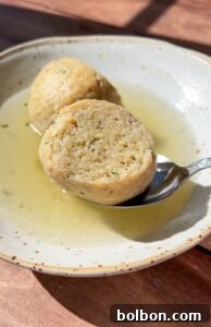 A bowl of clear broth with two sliced matzo balls, one resting on a spoon, sits on a wooden surface in bright sunlight—capturing the essence of Authentic Matzo Ball Soup.