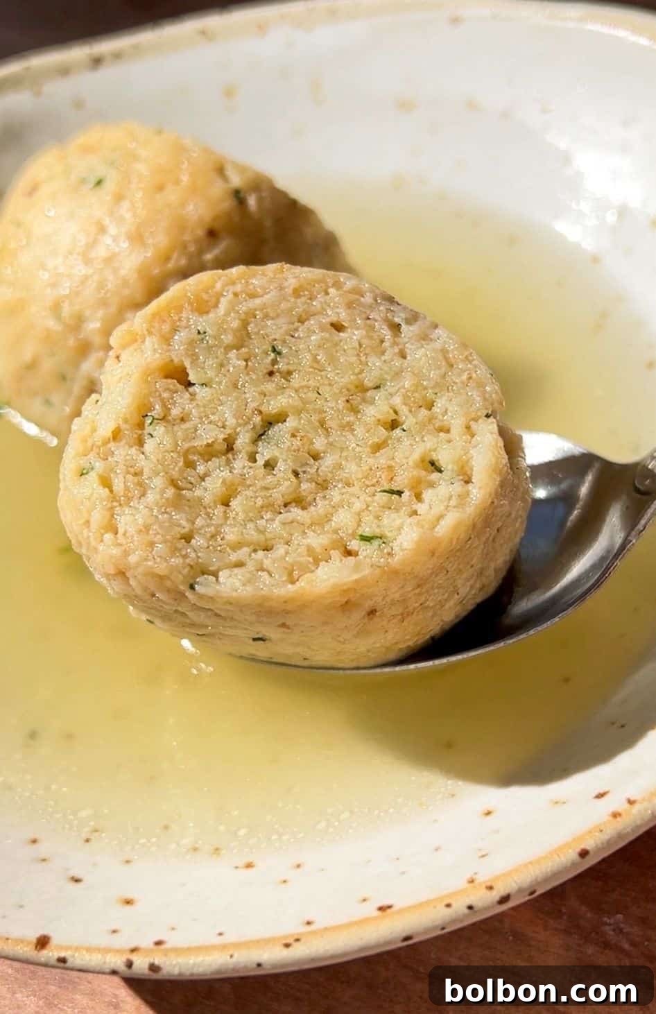 A close-up of Authentic Matzo Ball Soup shows two round matzo balls in clear broth, with one being lifted by a spoon from a shallow, speckled ceramic bowl.