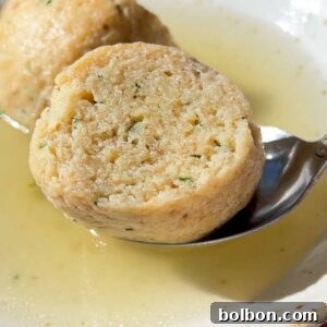 A close-up of a bowl of Authentic Matzo Ball Soup with clear broth and two matzo balls, one being lifted by a spoon. The fluffy texture and visible herbs highlight the traditional comfort of this classic dish.
