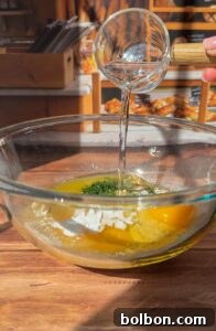 A hand pours water from a small glass cup into a large glass bowl containing eggs, flour, and herbs on a wooden countertop, preparing the base for Authentic Matzo Ball Soup in a cozy bakery setting.