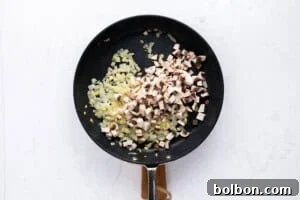 A black frying pan on a white surface contains sautéed chopped onions and freshly added chopped mushrooms, with both ingredients partially mixed.